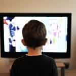 The back of a child sitting in front of and watching a television screen.