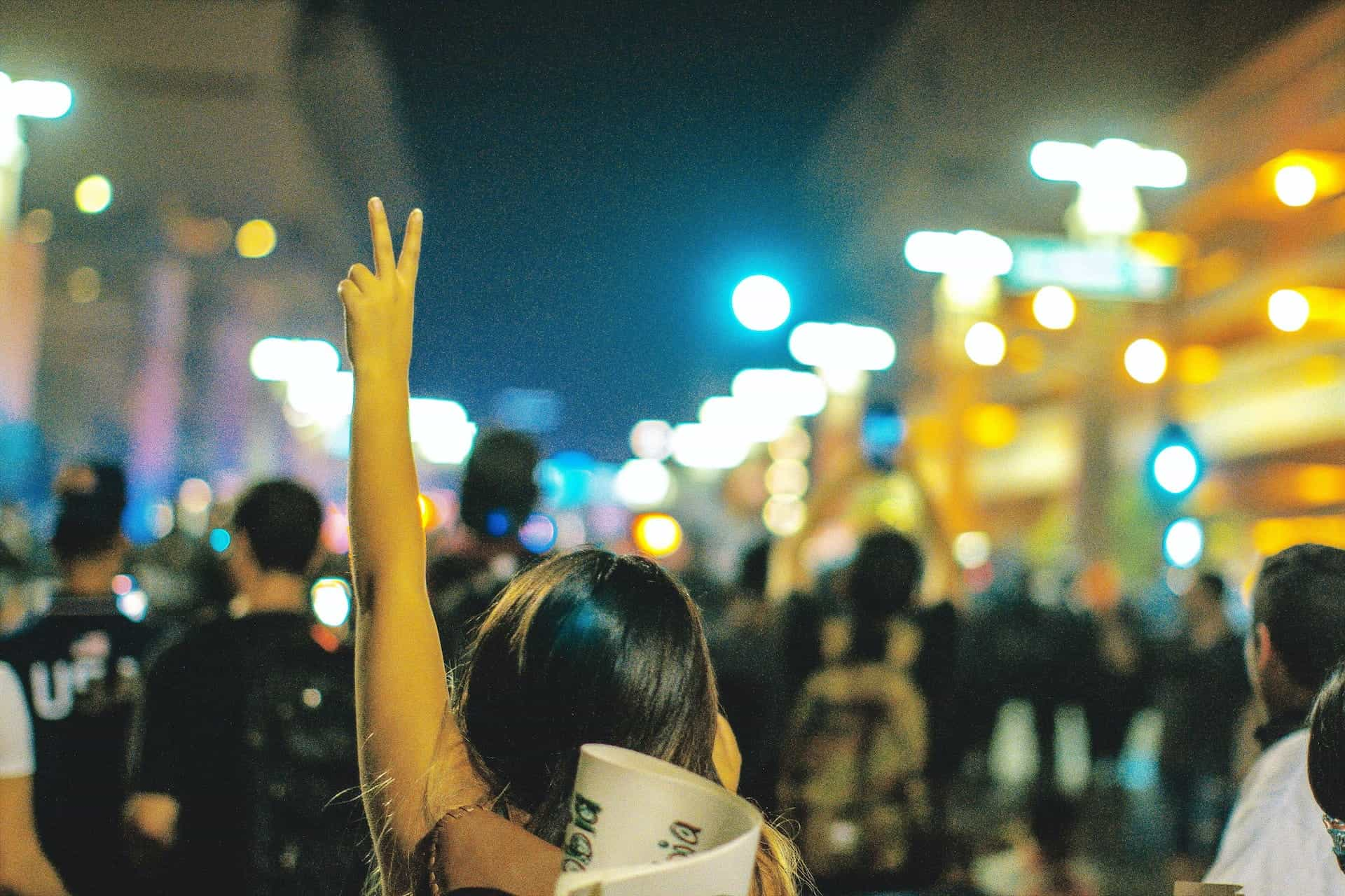 From behind, a woman raises her hand with the peace sign in a street protest at night.