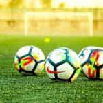 Three soccer balls sit on a green field with a net in the distance behind them.