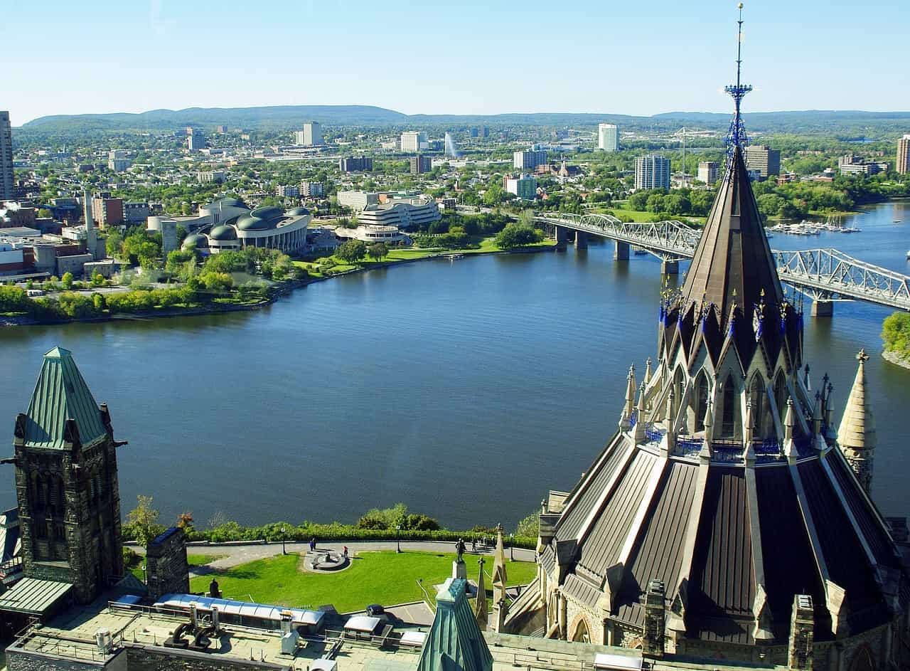 A view of Ottawa from the top of the Houses of Parliament in the city center, with a large river dividing the city in two and with several tall buildings sprawling out into the green landscape in the distance.