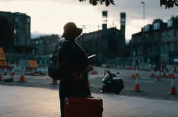 A traveler stands in a town square holding a red suitcase, backpack and hat